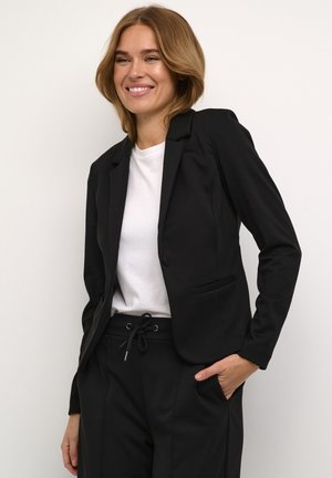 Černý blazer se strukturovanými klopami, zapínání na jeden knoflík a předními kapsami. Nosený přes bílou košili s černými kalhotami. Hladká textura látky.