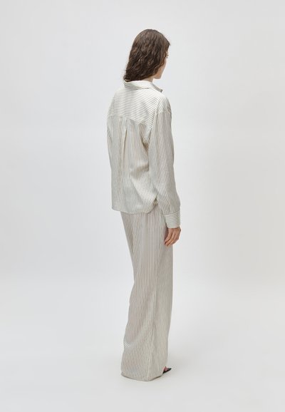 Göndör hajú nő oldalirányban áll, laza, fehér csíkos ingben és hozzá illő, bőszárú nadrágban, egyszínű háttér előtt.