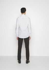 Cămașă albă cu mâneci lungi, cu un model subtil, cu manșete butonate și un tiv curbat, asociată cu pantaloni negri și pantofi, văzută din spate.