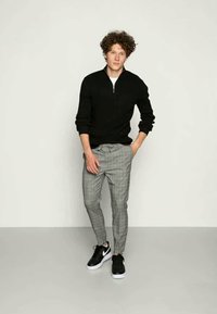 Casaco preto com fecho éclair, calças cinza xadrez ajustadas e ténis pretos. O conjunto apresenta um corte moderno, adequado para ambientes descontraídos.