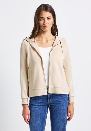 Haut de survêtement zippé beige clair avec un design ajusté, capuche à cordon de serrage et poignets côtelés. Comprend des poches latérales et des accents à rayures subtils.
