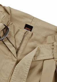 Beige bukser med D-ring bælte, med en metal lynlås, synlige syninger og et sort mærke med teksten "BOSS" på taljen.