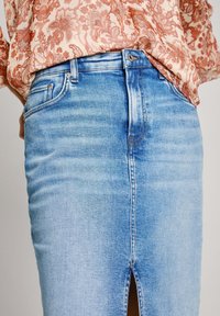Falda lápiz de mezclilla azul con una abertura frontal, con una cintura alta y desvanecido sutil. Combinada con una blusa de patrón floral claro.