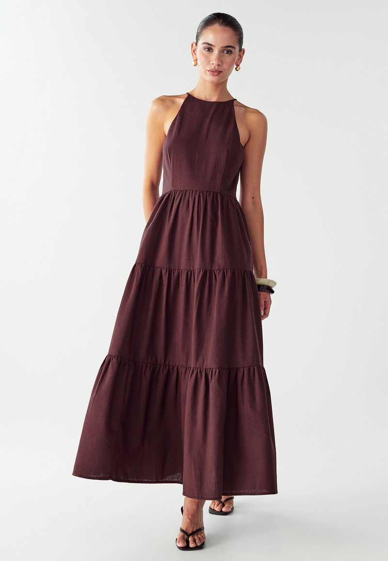 Nainen, joka on pukeutunut hihattomaan, kaula-aukoltaan halterneck maroon-maksimekkoon, jossa on kerroksellinen hame, mustiin sandaaleihin, kultaisiin korvakoruihin ja paksuihin rannekoruihin.