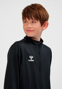 Svart zip-up-jacka i slätt tyg med hög krage och vit logotyp på vänster sida, bärs av ett barn med kort brunt hår.