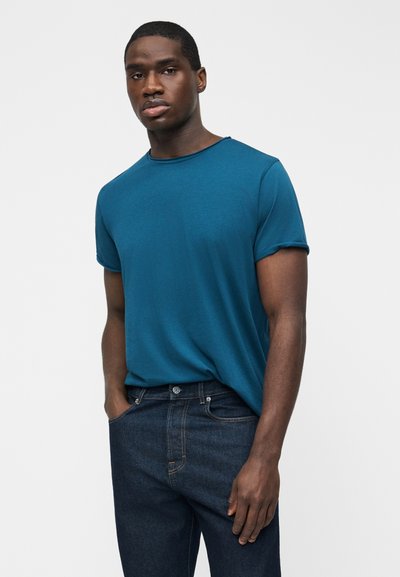 Μαύρος άνδρας στέκεται με τα χέρια στις τσέπες, φοράει μπλε κοντομάνικη μπλούζα και σκούρο μπλε τζιν σε απλό λευκό φόντο.