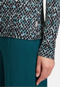 Langarmoberteil mit einem schwarz und türkisfarbenen geometrischen Muster und weißen Gitterlinien, kombiniert mit türkisfarbenen Hosen. Etikett auf der Seite des Oberteils sichtbar.