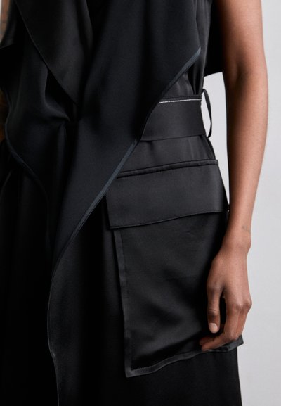 Black sleeveless garment with a flowing design, featuring a prominent pocket and contrasting stitching details along the edges.