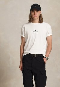 Νεαρός άνδρας φορεμένος με λευκό T-shirt Ralph Lauren, μαύρο cargo παντελόνι, μαύρο καπέλο και ρολόι χειρός, στέκεται με το ένα χέρι στην τσέπη.