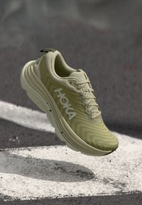 Hoka løpesko, lysegrønt meshoverdel, polstret såle, teksturert design, fremtredende logo på siden, og snøresystem foran.