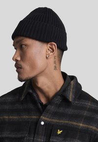 Beanie nera a costine indossata da una persona, abbinata a una camicia scozzese scura con accenti gialli e colletto button-down. Tatuaggio ben visibile sul collo.
