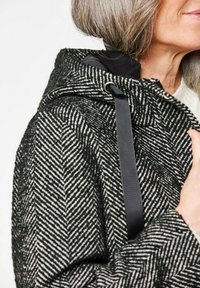 Zwart-witte herringbone-patroon jas met een gestructureerde stof, ritsluiting en een zwarte bandaccent op de kap.