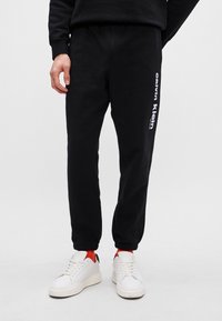 Juodos sportinės kelnės su elastiniu įsiuvimu, ant kairiojo kojos atvaizduojamas baltas 'Calvin Klein' logotipas. Dėvimos su baltais sportbačiais ir raudonomis detalėmis.