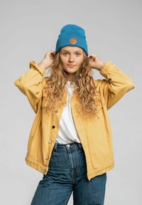 Żółta dżinsowa kurtka z srebrnymi guzikami, biała koszula pod spodem, niebieska czapka beanie z naszywką. Modelka ma długie, kręcone włosy ułożone swobodnie.