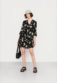 Vestido floral preto com um design evasê, decote em V, mangas 3/4 e camadas com babados. Combinado com um chapéu verde e sandálias pretas.