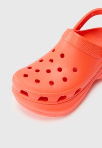 Oranžni plastični klomp z ventilacijskimi luknjami, vzorčasto pasom in zadnjim trakom. Gladka površina in zaobljena oblika. Strukturiran podplat.