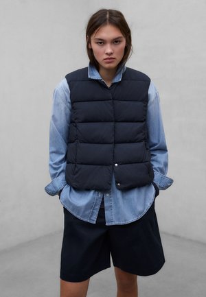 Giovane con espressione seria che indossa un gilet imbottito blu navy sopra una camicia di jeans azzurro chiaro e pantaloncini neri, in piedi davanti a uno sfondo grigio uniforme.