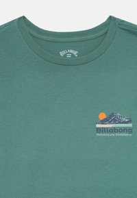T-shirt verde acqua con collo rotondo, logo Billabong e testo "Adventure Division", con grafica di una montagna e un sole arancione sul petto.