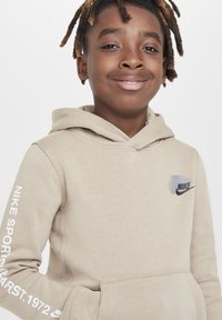 Hanorac bej Nike cu buzunar frontal tip kangaroo, glugă cu șnur și text alb pe mânecă. Dispune de un logo negru pe piept.