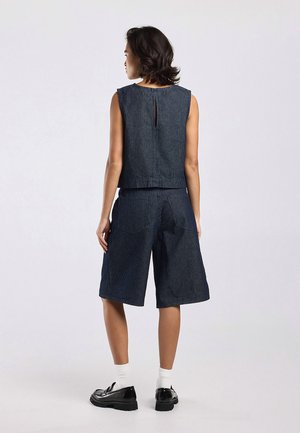 Femme de dos portant un haut sans manches rayé bleu marine et un short large assorti arrivant au genou, avec des chaussettes blanches et des chaussures noires.