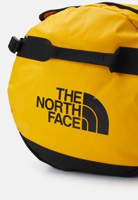 Žuta vodootporna torba s crnim logom The North Face, s zakrivljenim oblikom i čvrstim ručkama i remenom za sigurno nošenje.