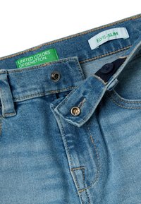 Jeans de mezclilla azul con un cierre a presión parcialmente desabrochado, etiqueta verde de "United Colors of Benetton" y etiqueta blanca de ajuste "Elvis-Slim" dentro de la cinturilla.