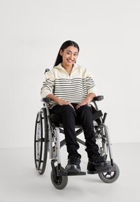 Maglione a righe, color crema e nero, con colletto a mezza zip, abbinato a pantaloni neri e sneaker sportive nere, indossato da una persona in sedia a rotelle.