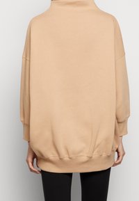 Beige oversized sweatshirt med hög krage, nedhasade axlar, ribbat avslut och mjuk textur. Slät tyg med minimal detaljering.