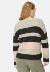 Vrouw met blond haar die een chunky gebreide trui draagt met brede zwarte, beige en roze horizontale strepen, en olijfgroene broek, met de rug naar ons toegekeerd.