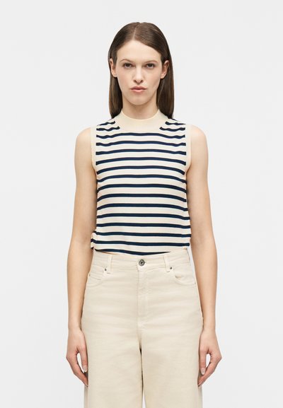 Femme portant un haut sans manches crème à rayures horizontales noires et un pantalon crème taille haute, debout devant un fond blanc uni.