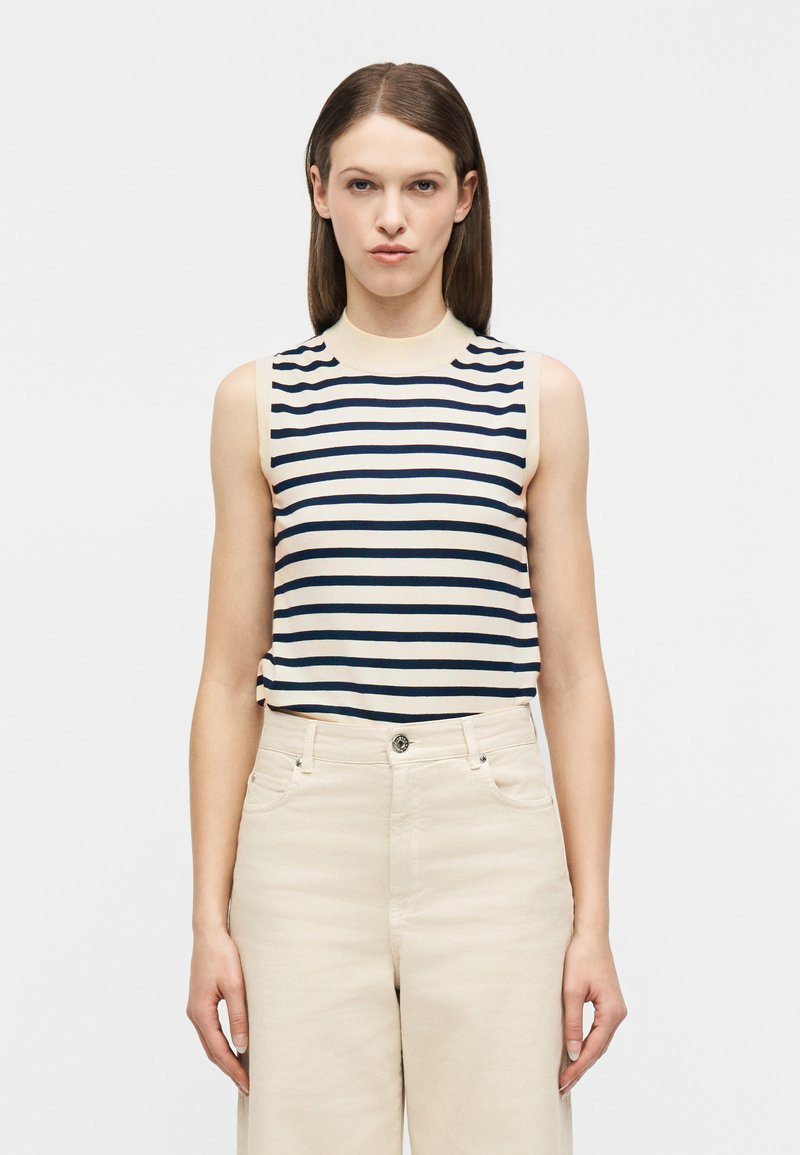 Γυναίκα που φοράει αμάνικη κρέμ μπ λούζα με μαύρες οριζόντιες ρίγες και ψηλόμεσο κρέμ παντελόνι, στέκεται μπροστά σε ένα απλό λευκό φόντο.