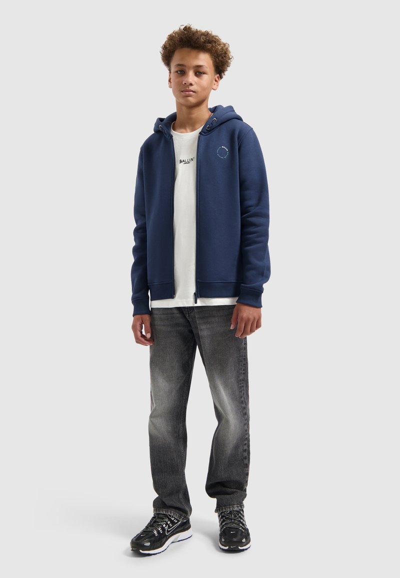 Marine kleurige hoodie met rits en trekkoorden, witte t-shirt met de tekst "BALLIN'", grijze jeans en zwarte sportschoenen. Krullend haar, neutrale achtergrond.