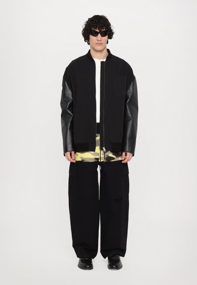 Jachetă bomber neagră cu mâneci din piele, tricou alb, pantaloni cargo cu model de camuflaj închis, pantofi negri și ochelari de soare întunecați.