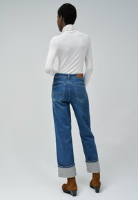 Plave traperice visokog struka, ravnog kroja, s rukavima na kraju nogavica, uparene s bijelim dugim rukavima i turtleneckom. Vidljive smeđe gležnjače.