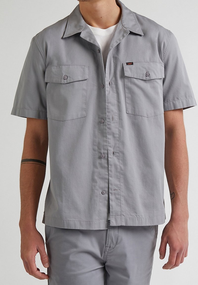 Camicia utility grigia a maniche corte in cotone. Presenta due tasche frontali, chiusura con bottoni e colletto a punta con vestibilità rilassata.