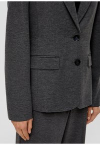 Blazer gris à deux boutons en tissu texturé. Dispose d'un revers cranté, de deux poches avant et d'une coupe ajustée.
