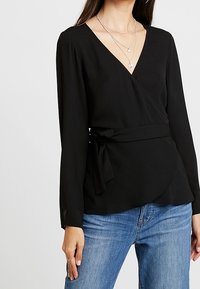 Sort omslagsbluse med lange ærmer, V-udskæring og bundet talje. Blødt stof med glat tekstur og flydende design. Bruges sammen med blå jeans.