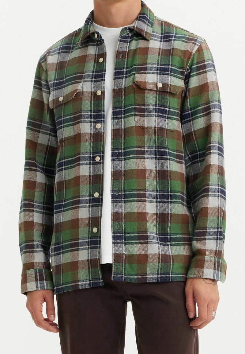 Homme portant une chemise à carreaux verte, marron, marine et blanche par-dessus un t-shirt blanc avec un pantalon marron foncé, debout, les bras détendus.