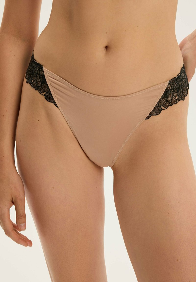 Beige Tang mit schwarzem floralen Spitzenbesatz an den Hüften. Weicher, glatter Stoff mit nahtlosem Design. Verstellbare Träger für individuelle Anpassung.