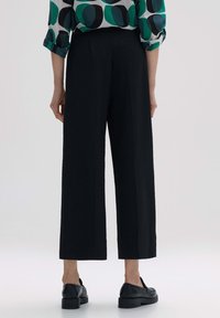 Pantalons larges noirs en tissu texturé. Conception taille haute avec une ceinture lisse. Portés avec une chemise à motifs verts et blancs.