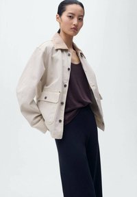 Veste oversize beige clair avec un col beige, des boutons foncés et des poches avant, portée sur un débardeur noir et un pantalon noir.