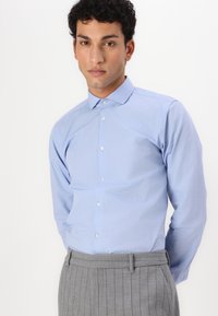 Camisa de botones azul claro con cuello de punta y mangas largas, hecha de un tejido suave. Llevada con pantalones grises a rayas.