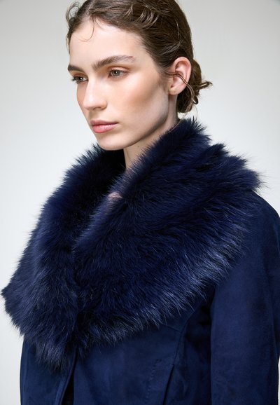 Femme à la peau claire et aux cheveux bruns attachés porte un manteau bleu foncé avec un grand col en fourrure duveteuse, regardant légèrement vers le bas et sur le côté.