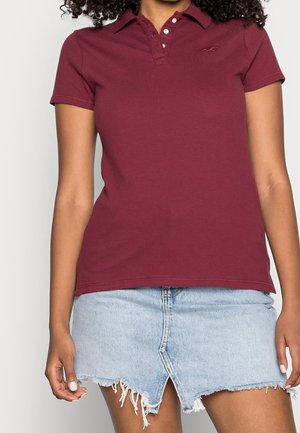 Burgundowa koszulka polo wykonana z bawełny z kołnierzykiem i trzema guzikami, w połączeniu z jasnoniebieską, postrzępioną spódnicą dżinsową.