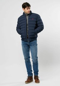 Hombre vestido con una chaqueta acolchada azul marino, un suéter de cuello de tortuga gris, jeans azules y botas de cordones marrones, de pie con las manos en los bolsillos de la chaqueta.