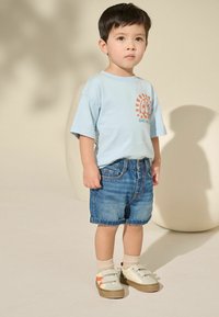 Helles blaues T-Shirt mit orangefarbenem Sonnenmotiv, Jeansshorts und beigen Sneakern mit zwei Klettverschlüssen, getragen mit hellen Socken.