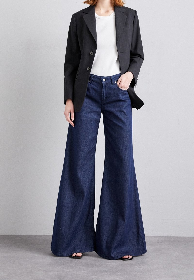 Femme portant un jean large foncé, un blazer noir, un haut blanc et des chaussures noires à bouts ouverts, debout contre un mur gris uni.