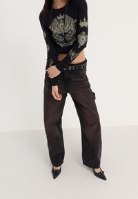 Black long-sleeve crop top with gold graphic designs, paired with loose dark brown pants and a black belt featuring metal eyelets.