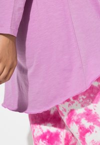 Violetti pitkähihainen paita pyöreällä helmalla ja pehmeällä tekstuurilla, yhdistetty valkoisiin leggingsseihin, joissa on vaaleanpunaiset batikkikuvioinnit ja näkyvää ompelua.