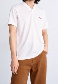 Bílé polo tričko s krátkým rukávem vyrobené z měkké látky, s nízkým výstřihem do V a malým červeným logem na levé hrudi, doplněné hnědými kalhotami.